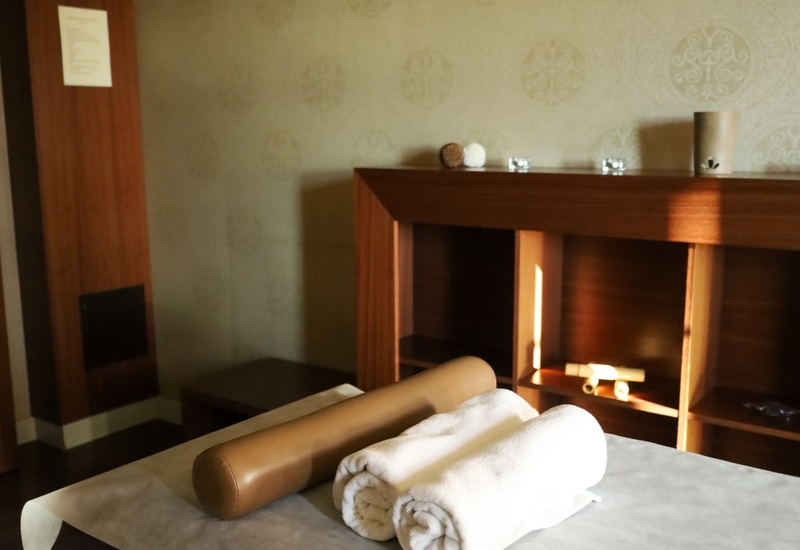 Pokój spa z leżanką do masażu, zwiniętymi białymi ręcznikami i wałkiem, drewnianymi półkami i delikatnie wzorzystą tapetą.