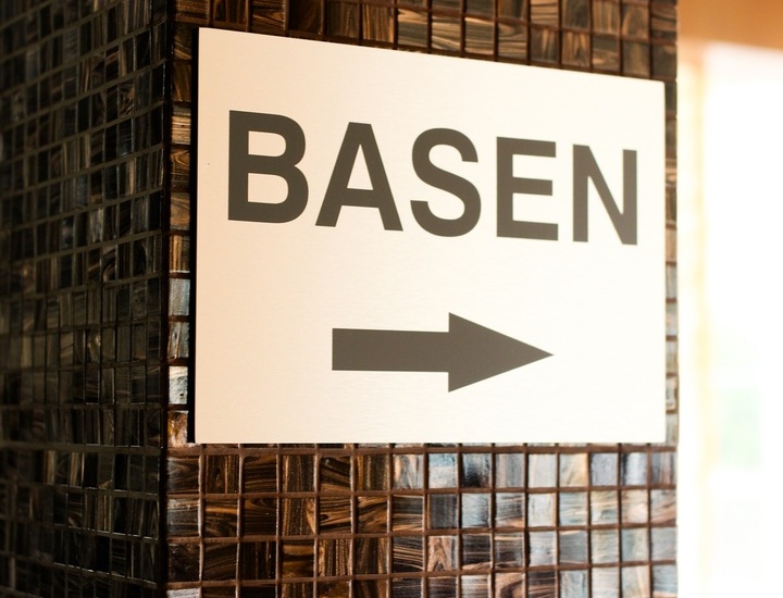 Znak 'BASEN' ze strzałką wskazującą drogę do basenu, przymocowany do kolumny w ciemnej mozaice.