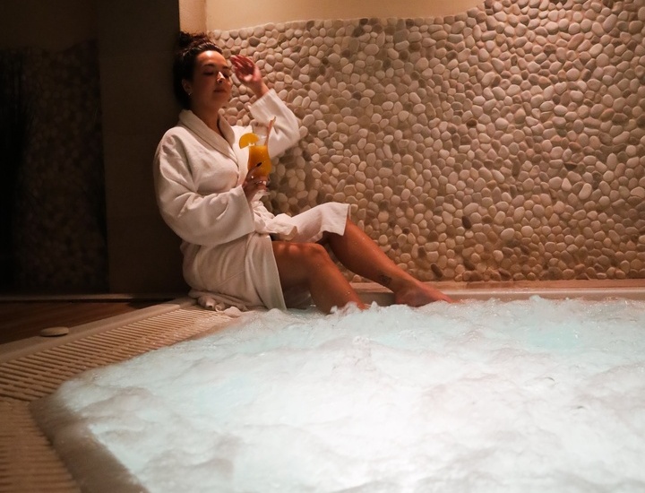 Gość w białym szlafroku relaksuje przy bąbelkowym jacuzzi, trzyma koktajl przy kamiennej ścianie w ciepłym świetle