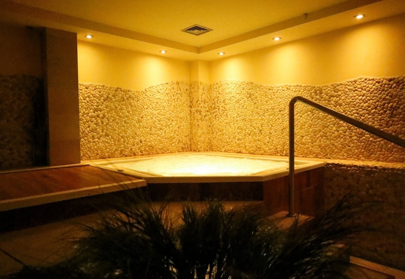 Jacuzzi w spa z ciepłym oświetleniem, ścianami wyłożonymi kamykami, drewnianym tarasem i metalową poręczą.