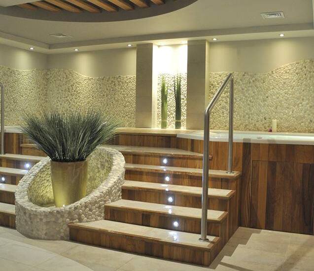 Spa wewnętrzne z podwyższonymi drewnianymi schodami prowadzącymi do wanny z hydromasażem, ściany z kamyków, nastrojowe oświetlenie i rośliny.