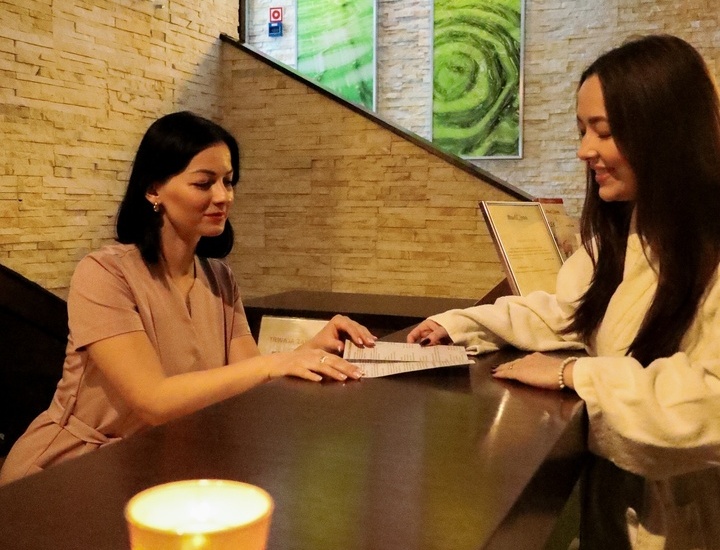 Przytulna recepcja spa, recepcjonistka obsługuje gościa w szlafroku przy kamiennym blacie, świece i zielone grafiki