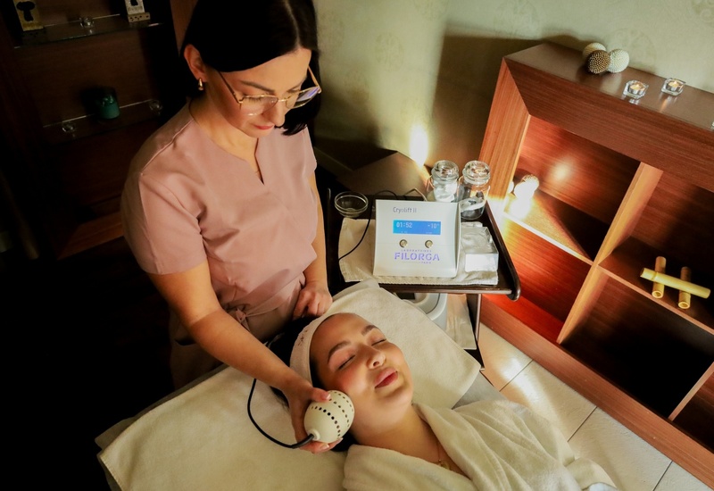 Kosmetyczka używa ręcznego urządzenia do twarzy na zrelaksowanej klientce w szlafroku na łóżku spa, drewniane półki i świeczki.