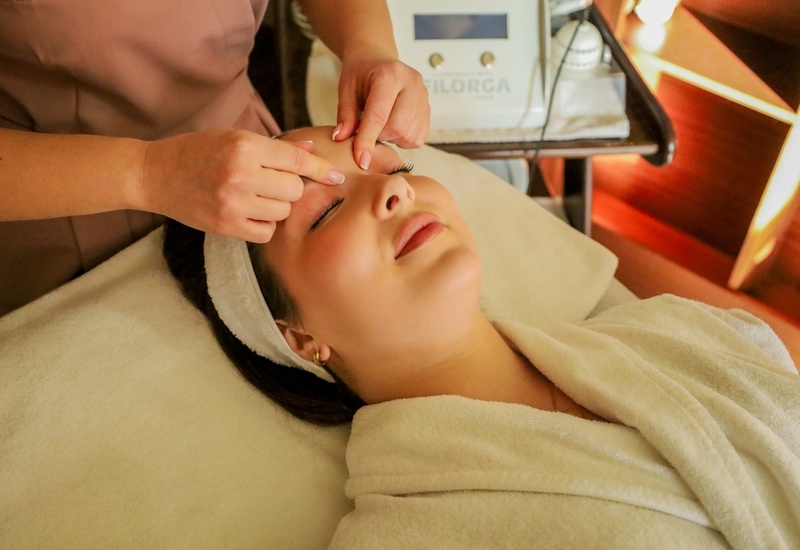 Gość w szlafroku otrzymuje relaksujący zabieg na twarz od kosmetyczki na łóżku spa, opaska na włosach, ciepłe światło.