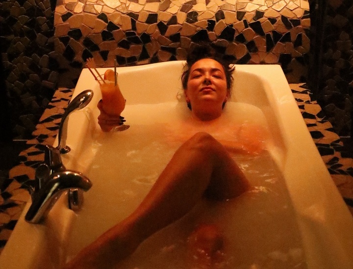 Gość relaksuje się w wannie spa z mozaikową ścianą przy świecach, trzyma koktajl.