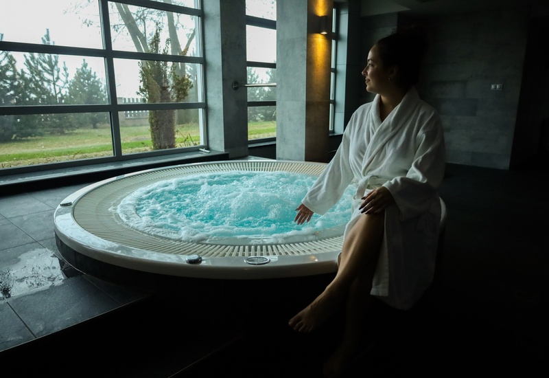 Kobieta w białym szlafroku siedzi przy bąbelkowym jacuzzi w spa, duże okna z widokiem na drzewa i ogród