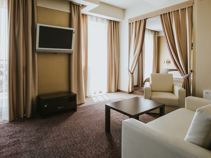 Priestranný hotelový apartmán s béžovou sedačkou a kreslom, konferenčným stolíkom, televízorom na stene, balkónovými dverami a oddelenou spálňou za závesom