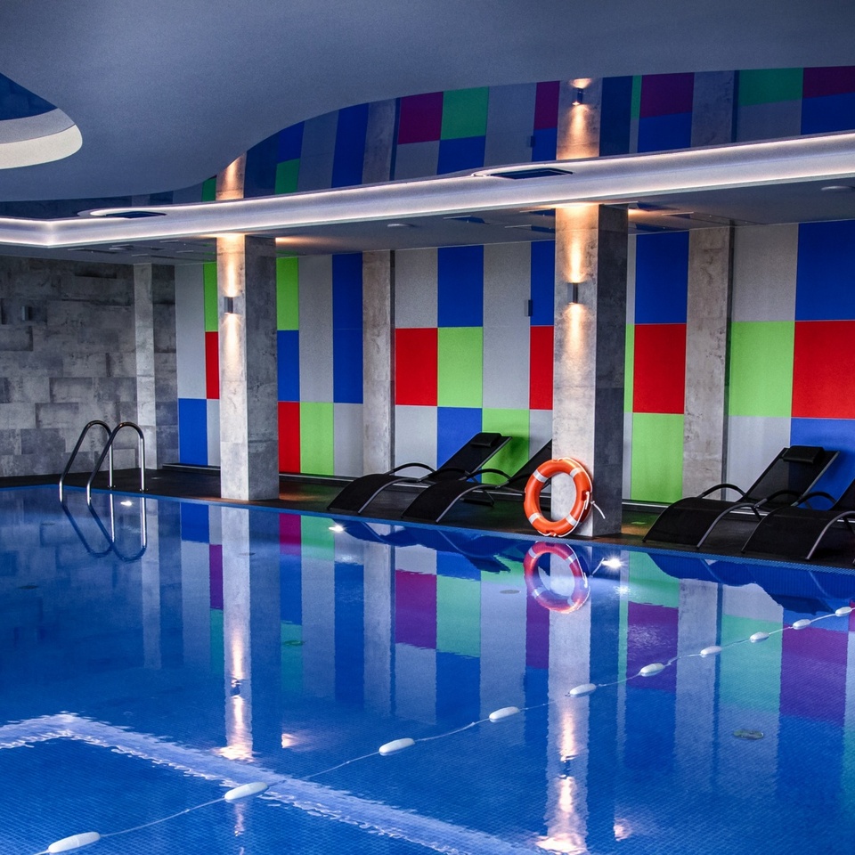Indoor hotel pool with blue tile, loungers, colorful geometric wall panels, soft ceiling lighting and life ring
