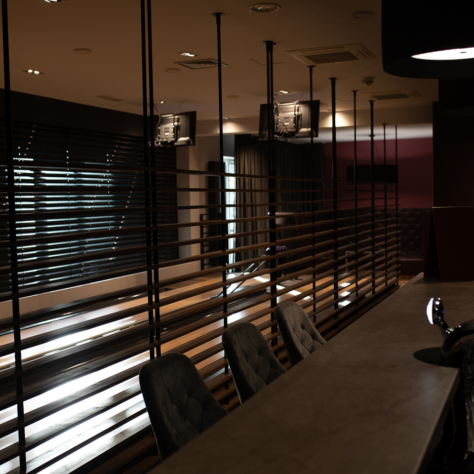 Moderne Hotelbar mit gepolsterten Hockern, Zapfhahn und Gläsern, Holzlamellen-Trennwand zeigt gedämpften Lounge-Bereich mit Fernsehern.