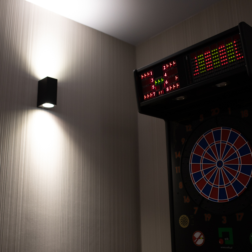 Elektronische Dartscheibe mit beleuchtetem LED-Display neben einem Wandleuchter auf strukturierter Tapete in einer Spielzone