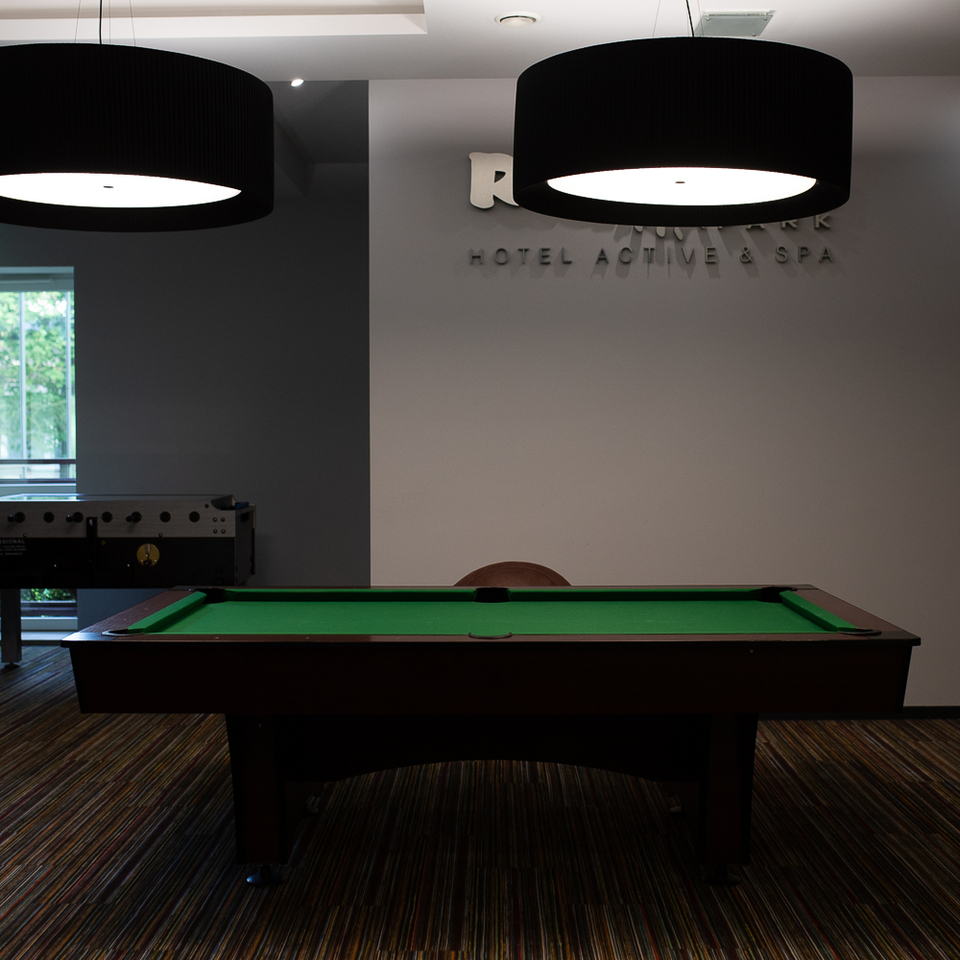 Hotel-Spielraum mit grünem Billardtisch unter zwei großen Pendelleuchten, Kickertisch, Sesseln und gestreiftem Teppich.