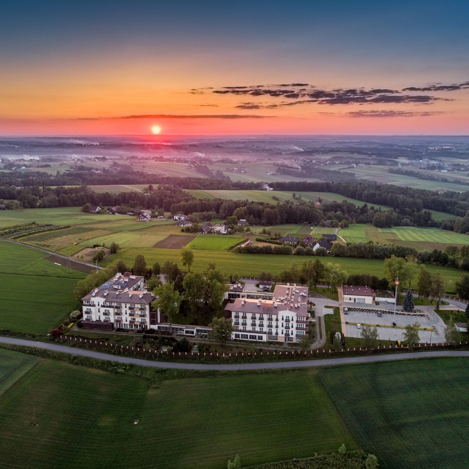 Pohled z ptačí perspektivy na venkovský hotel s balkony, parkovištěm a zahradami mezi zvlněnými poli za východu slunce