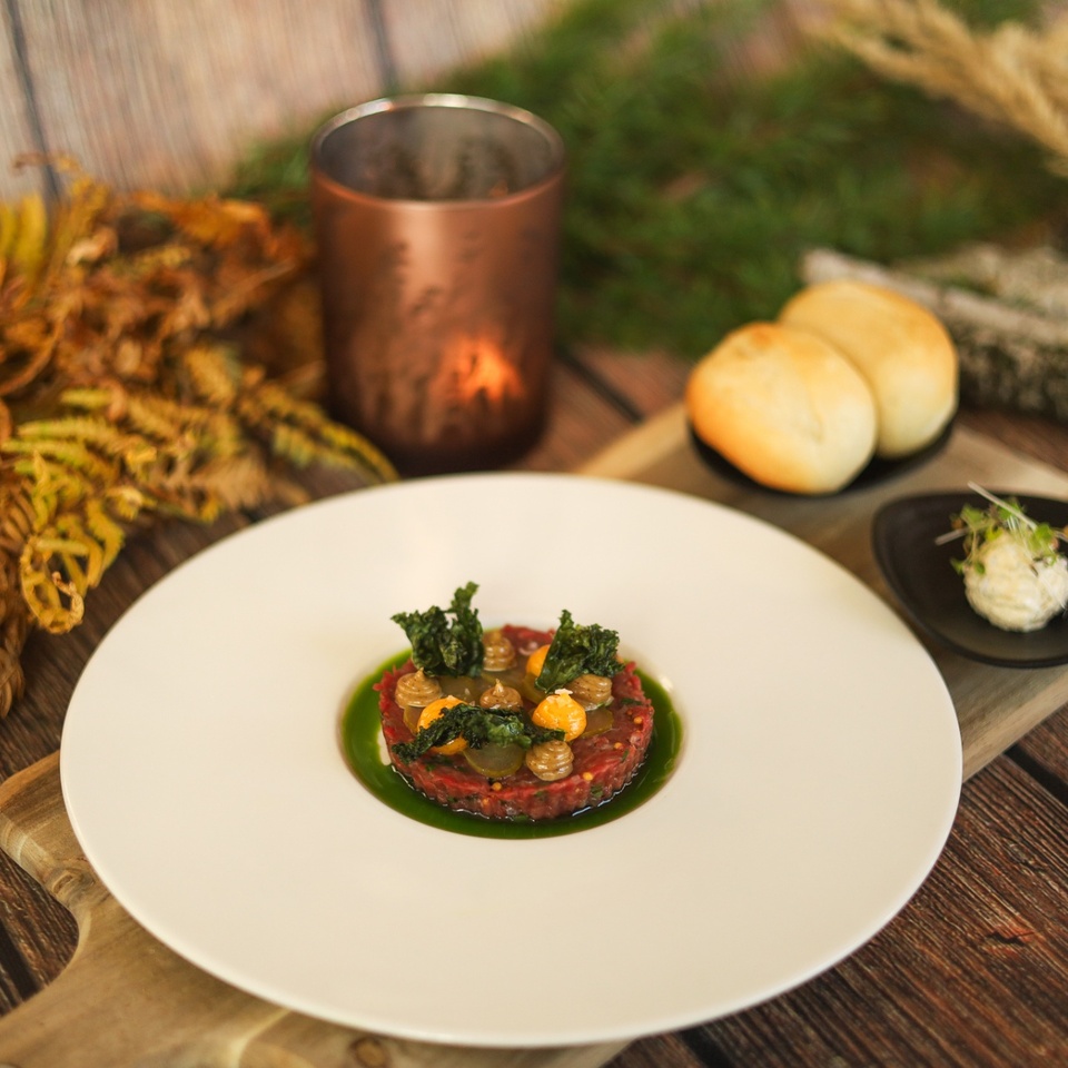 Hovädzí tatarák s chrumkajúcim kelom a bylinným olejom, podávaný s žemľami a maslom na rustikálnej doske pri sviečke