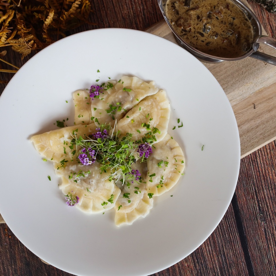 Domáce pirohy s mikrozeleninou a fialovými jedlými kvetmi, podávané s krémovou hríbovou omáčkou v sosjerke na drevenej doske.