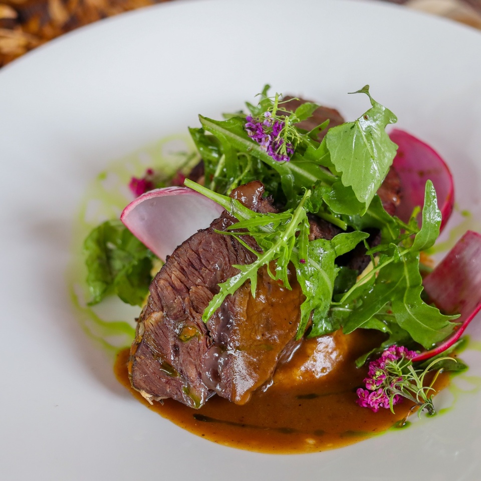 Plátky šťavnatej hovädziny v tmavej omáčke, rukola, plátky reďkovky a jedlé kvety na bielom tanieri