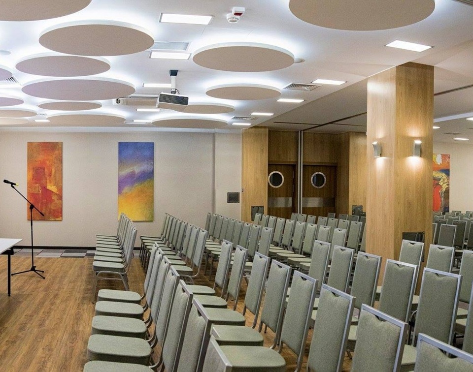 Moderná konferenčná sála s radmi stoličiek, obrazovkou, stolíkom pre prednášajúceho, okrúhlymi panelmi a drevenými stĺpmi