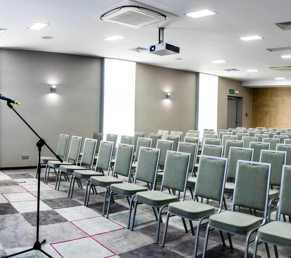 Moderná konferenčná sála v hoteli so sadou zelených stoličiek, projektorom a obrazovkou, stolom s laptopom a mikrofónmi, kobercom a jasným osvetlením