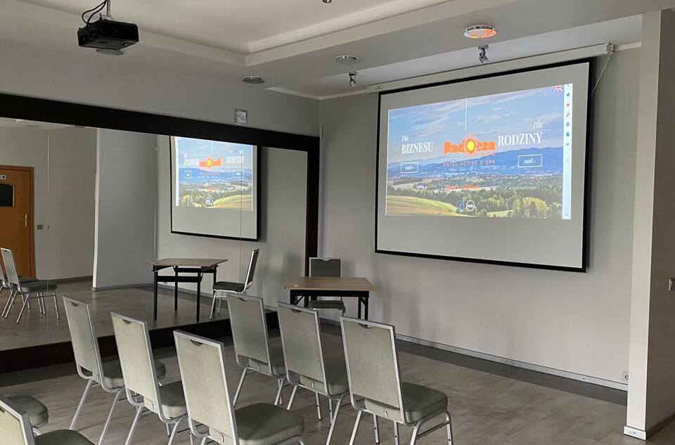 Malá konferenčná sála v hoteli s projektorom na strope a veľkou obrazovkou, radmi čalúnených stoličiek, zrkadlom a stolíkmi.