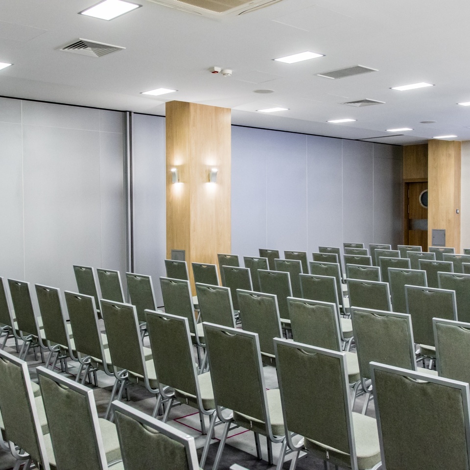 Konferenčná sála v hoteli s radmi zelených stoličiek, projektorom, mikrofónom, drevenými stĺpmi a jasným osvetlením