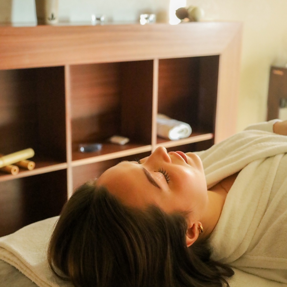 Žena v župane odpočíva na spa lehátku so zavretými očami, teplé osvetlenie, drevené police a zrolované uteráky.