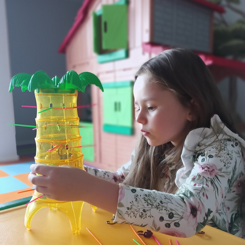 Mädchen konzentriert beim Spiel: baut gelben Palmen-Turm, steckt bunte Stäbchen und Kunststofffiguren an einem Spieltisch zusammen.