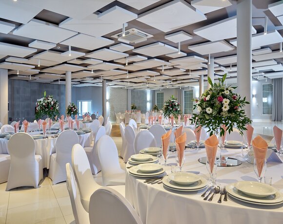 Elegancka sala bankietowa na wesele z okrągłymi stołami, białymi pokrowcami, brzoskwiniowymi serwetkami, kwiatowymi dekoracjami i nowoczesnym sufitem