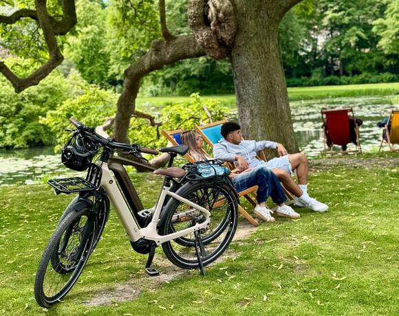 Paar entspannt in Liegestühlen am See unter großem schattenspendendem Baum, elektrische Hotelräder geparkt.