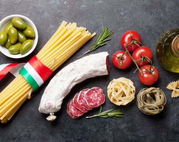 Włoskie składniki do potraw: spaghetti z trójkolorową wstążką, pokrojone salami, pomidory na gałązce, oliwki, oliwa i rozmaryn