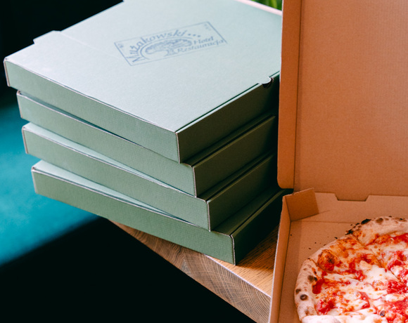 Otwarte pudełko z pizzą margherita na drewnianym stole, obok stos zielonych pudełek i wazon z różowymi tulipanami