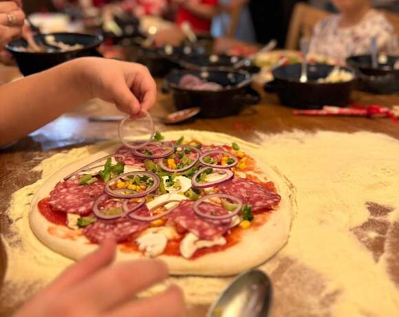 Dzieci przygotowujące pizze przy wspólnym stole — salami, czerwona cebula, kukurydza i świeże dodatki