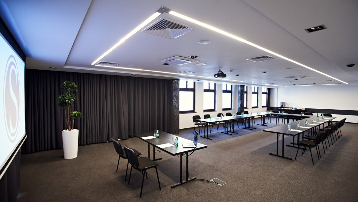 Large conference room with U-shaped tables, black chairs, projector screen, bottled water at each seat.
