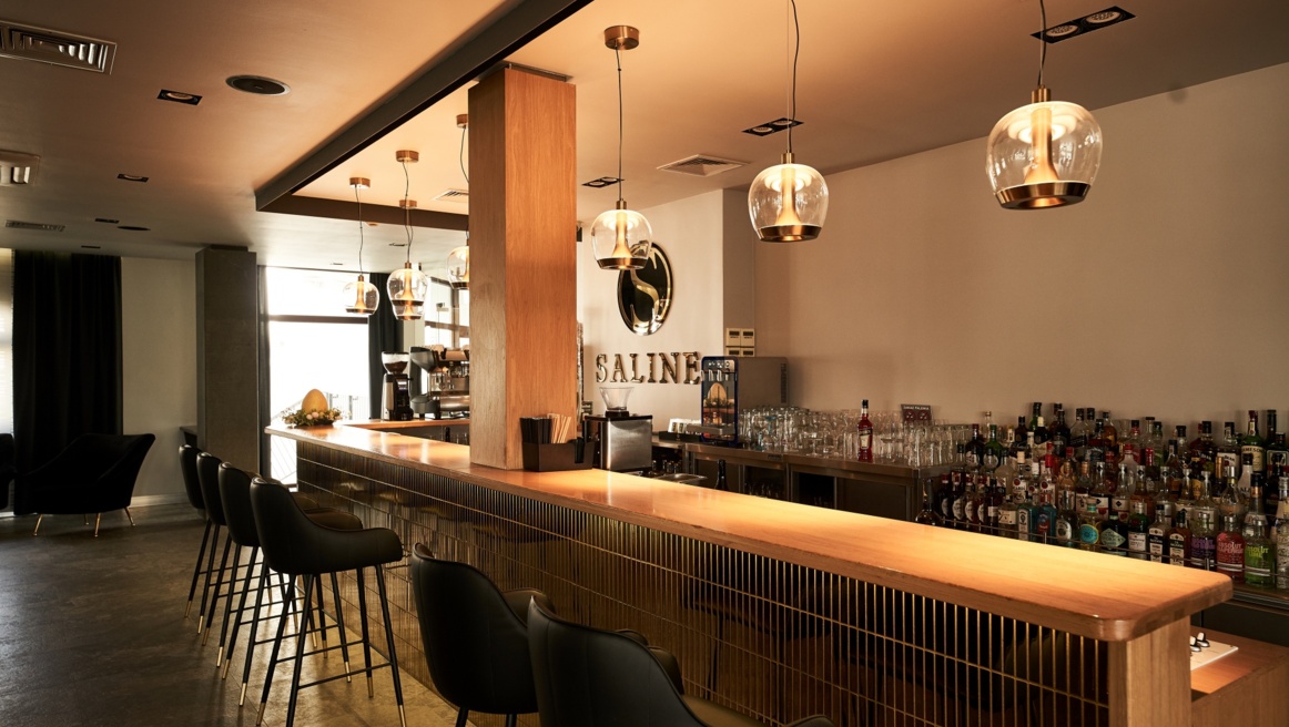 Cozy hotel bar with wooden counter, black bar stools, hanging glass pendants, stocked liquor shelves.