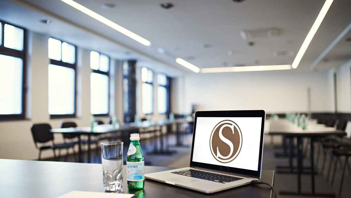Laptop displaying circular 'S' logo on table, glass and water bottle beside it, blurred conference room background.