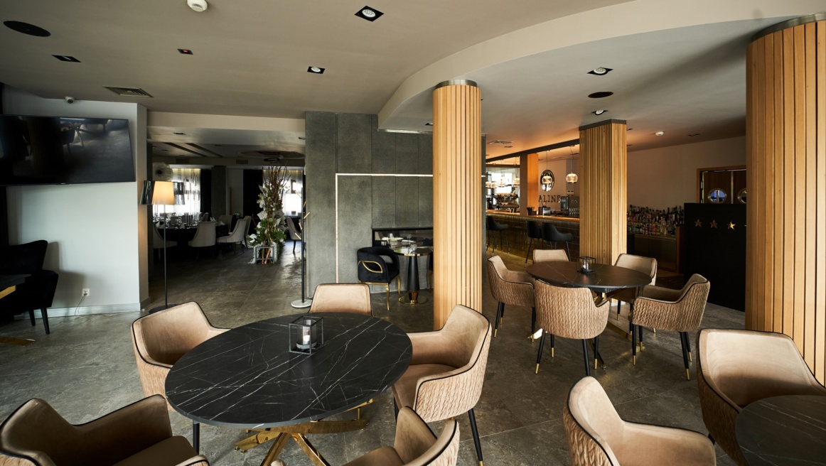 Warm lounge area with round black tables, cushioned tan chairs, wooden columns and ambient bar lighting.