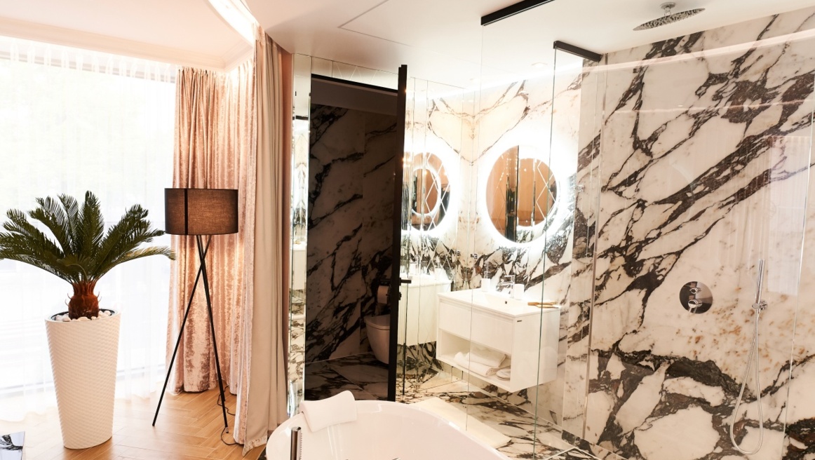 Luxurious marble bathroom with freestanding bathtub, glass shower, illuminated round mirrors, potted plant and floor lamp.