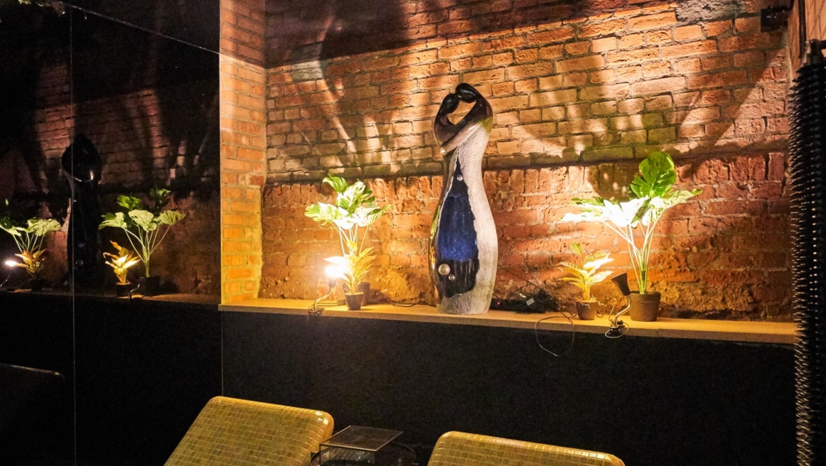 Ambient corner with illuminated abstract sculpture, potted plants, exposed brick and dramatic shadows.