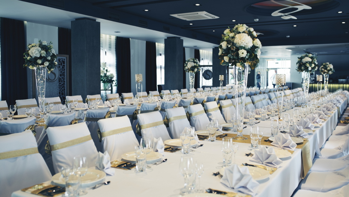 Bright banquet hall with long white-and-gold tables, neatly folded napkins, tall white floral vases.