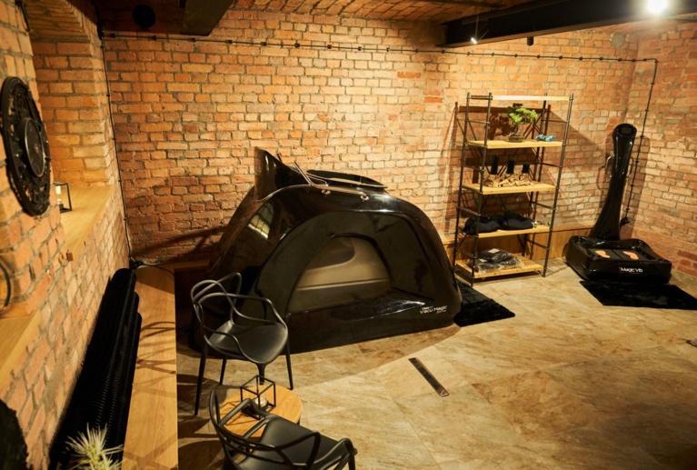 Industrial-style wellness room showing a black pod machine, vibration platform, wooden shelving and tiled floor.