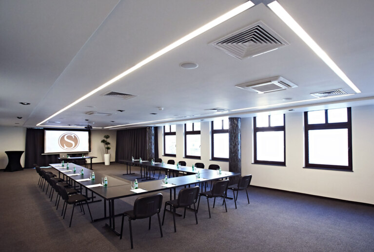 Spacious conference room with U-shaped tables, chairs, projector screen and bottled water, bright daylight windows.