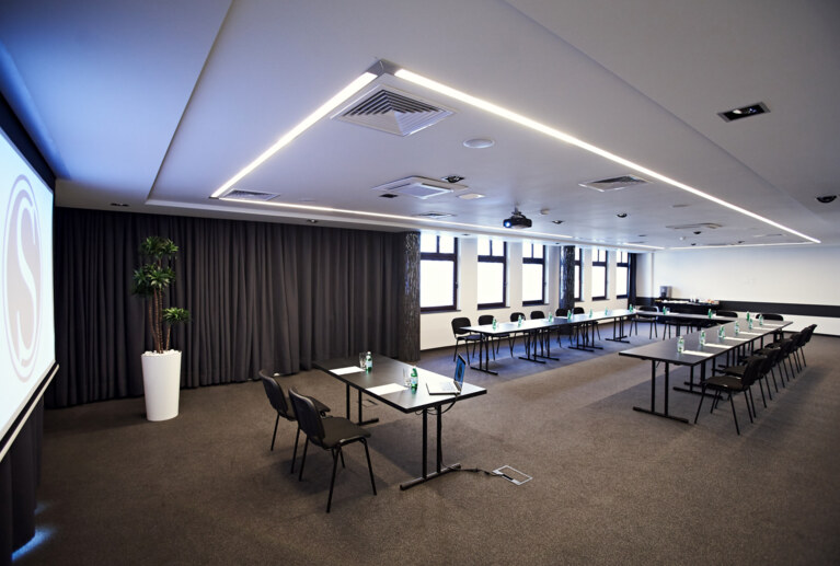 Spacious training room setup with arranged chairs, projection screen, and floor plant by dark curtains.