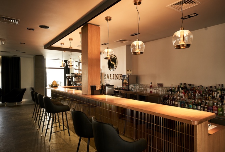 Modern bar showing polished counter, backlit liquor display, 'SALINE' logo on wall, soft lighting.