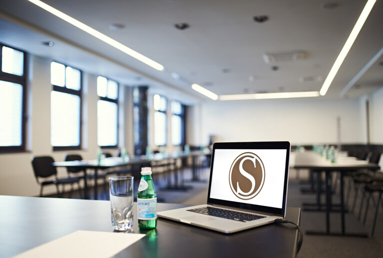 Close-up laptop and stationery on conference table, bottled water and rows of empty chairs receding into background.