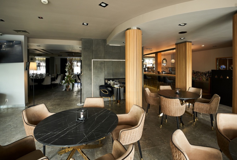 Elegant lounge corner showing black stone tables, plush tan chairs, view into dining area and decorative plants.