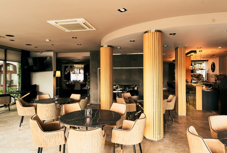 Empty dining lounge arranged with circular tables, plush chairs, floor lamp, and decorative wood columns.