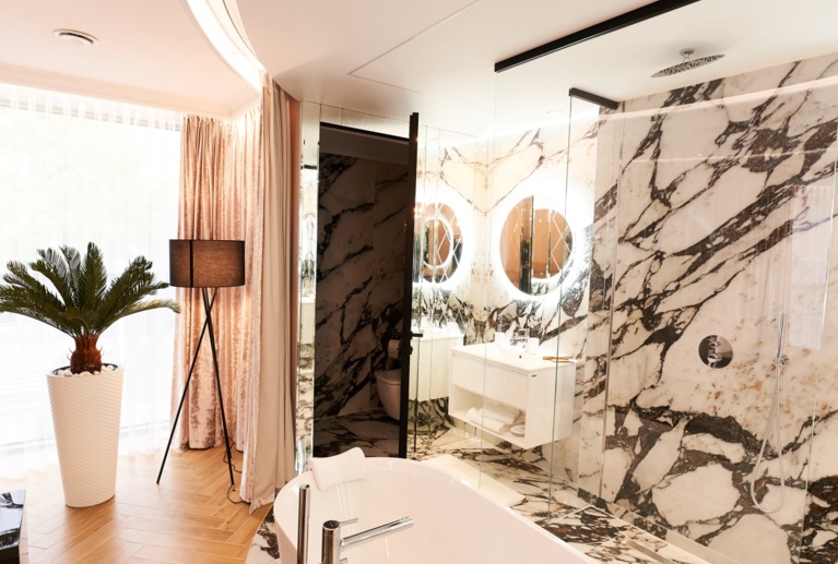 Modern vanity reflected in lighted round mirrors, marble walls, glass shower, freestanding tub foreground.