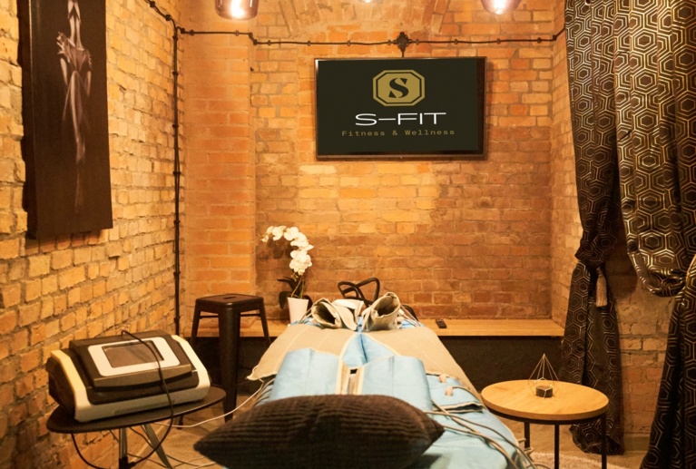 Warmly lit treatment room with exposed brick, decorative curtain, stool, and wellness equipment.