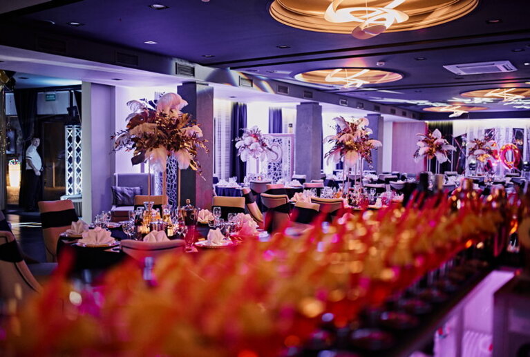Purple-lit banquet hall with blurred row of glasses in foreground, tables set, tall floral centerpieces.