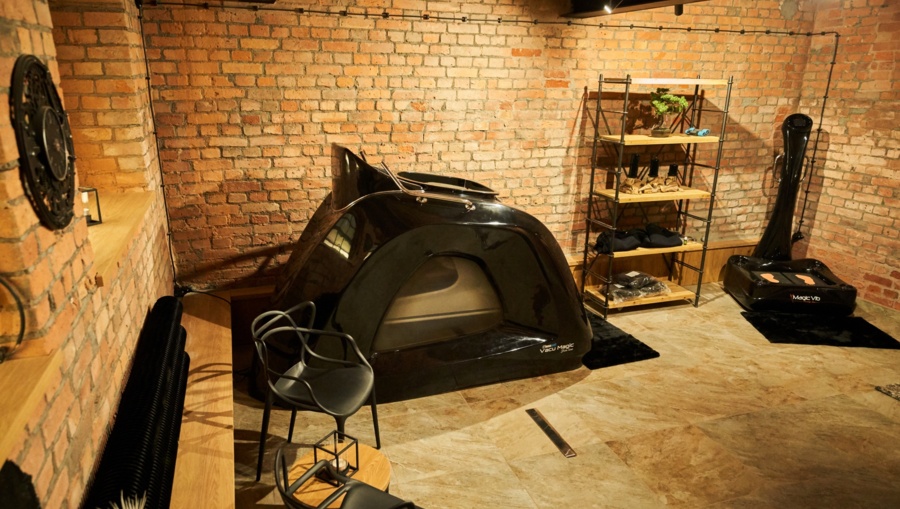 Brick-walled treatment room with black spa capsule, chairs, shelving and ambient warm lighting.
