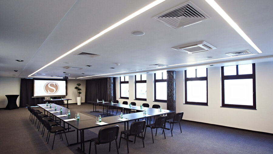 Well-lit meeting room with stacked chairs, tables arranged for presentation, clean minimalist decor.