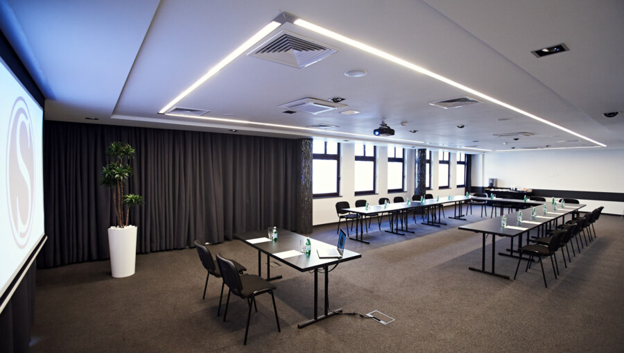 Meeting space with long tables, notepads, laptop on front table, neutral carpeting, bright ceiling lights.